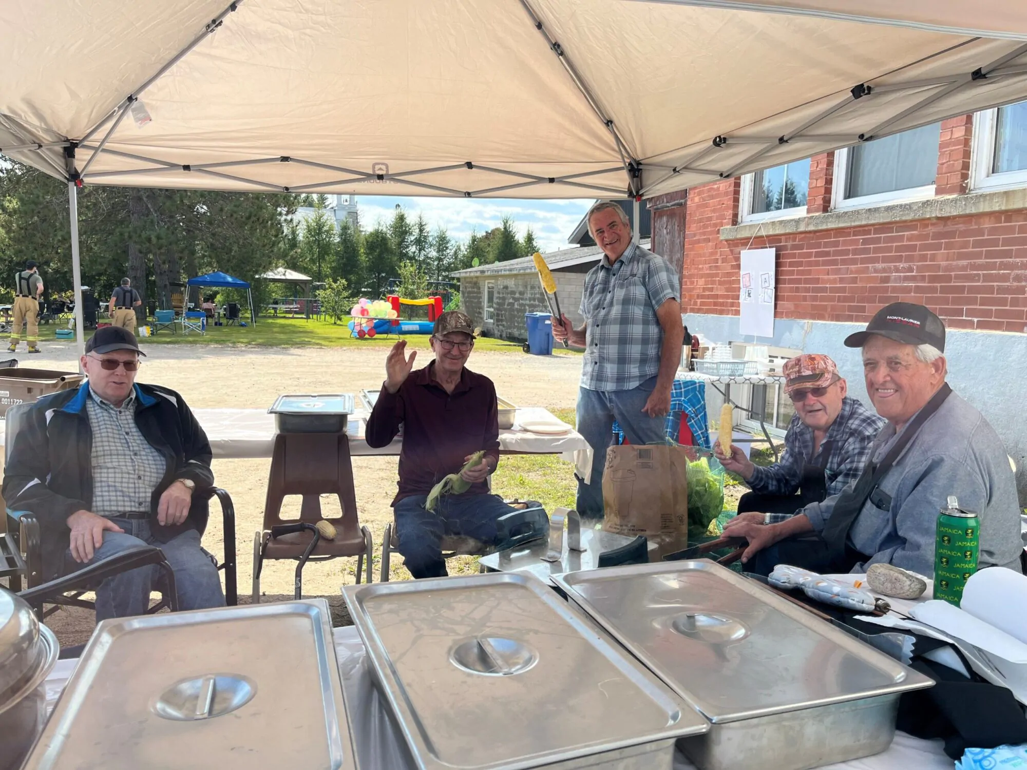 Un groupe d’hommes assis et debout sous un chapiteau, préparant et servant du maïs dans une ambiance conviviale. À l’arrière-plan, on aperçoit des installations extérieures et des activités festives.