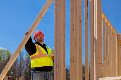 Un travailleur avec un dossard jaune et un casque rouge inspecte un mur en charpente en construction sur un chantier.