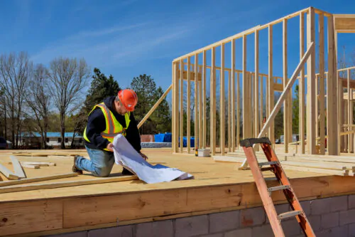 Un travailleur de la construction regarde des plans, agenouillé sur un plancher de bois d'oeuvre, près d'un mur en train d'être érigé, sur un chantier.