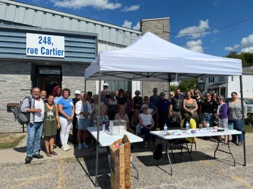 Un groupe de personnes réunies à l’extérieur d’un bâtiment situé au 248, rue Cartier, sous un chapiteau blanc et autour de tables installées pour l’occasion.