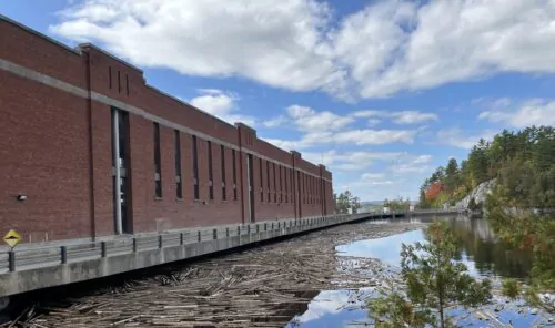 Un long bâtiment de briques rouges d'un barrage hydroélectrique devant lequel se trouve un passage pour les voitures, avec un plan d'eau et des billots de vois qui flottent au premier plan.