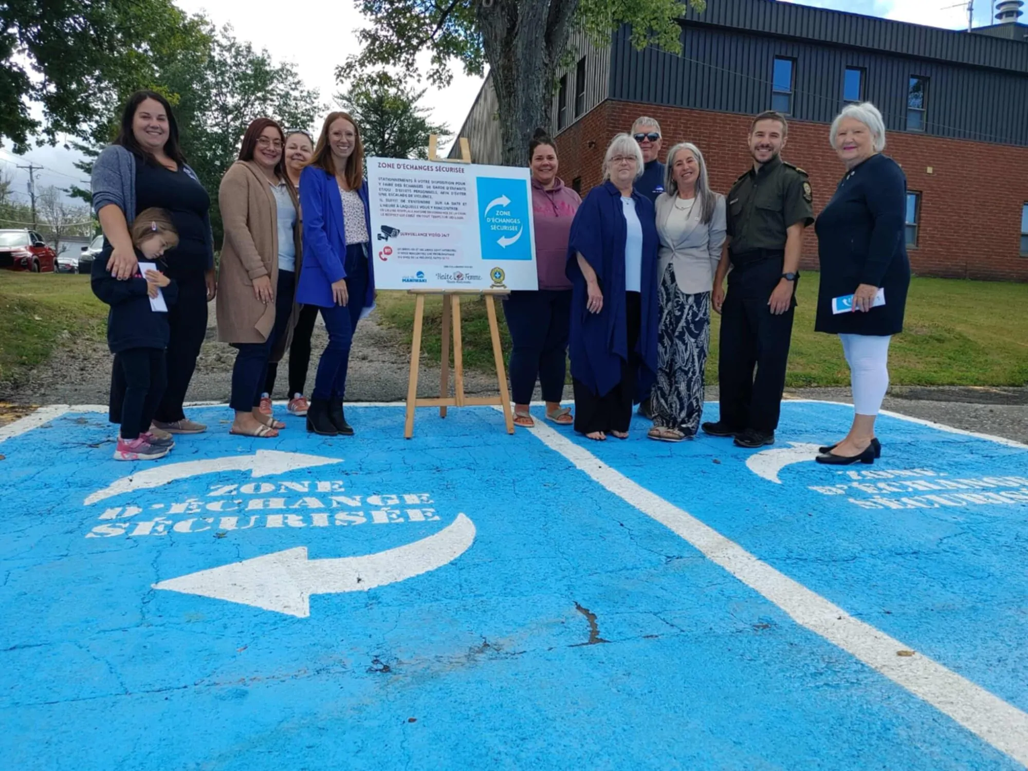 Un groupe de personnes posant à l’extérieur sur un espace de stationnement peint en bleu. Au centre, un panneau annonce la création d’une « zone d’échange sécurisée », un endroit destiné à permettre des échanges entre particuliers dans un lieu sûr. L’arrière-plan montre un bâtiment en brique et des arbres.