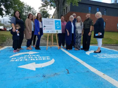 Un groupe de personnes posant à l’extérieur sur un espace de stationnement peint en bleu. Au centre, un panneau annonce la création d’une « zone d’échange sécurisée », un endroit destiné à permettre des échanges entre particuliers dans un lieu sûr. L’arrière-plan montre un bâtiment en brique et des arbres.