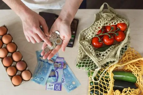 Une personne compte de la monnaie et des billets canadiens au-dessus d’une table, à côté d'aliments frais comme des œufs, des tomates et des légumes dans des sacs réutilisables.
