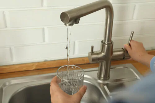 Une femme fait couler de l'eau d'un robinet de cuisine dans un verre d'eau qu'elle tient dans la main au-dessus d'un évier.