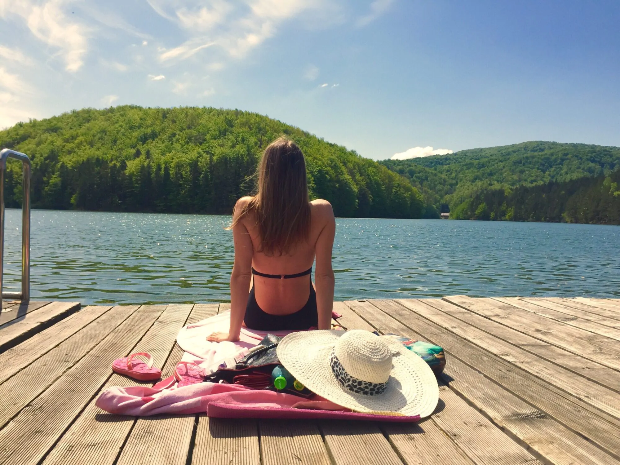 Une femme en maillot de bain assise sur un quai avec des accessoires estivaux regarde un lac.