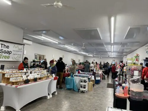 On voit une grande salle intérieure animée, remplie de kiosques de vente et de nombreux visiteurs. Il s’agit d’un marché ou d’une vente de Noël, où des artisans et commerçants locaux proposent des produits variés (savons, décorations, objets cadeaux). L’ambiance est chaleureuse et festive, avec beaucoup d’échanges entre exposants et clients.