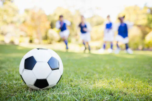 Vue de proche d'un ballon de soccer posé sur une pelouse avec quatre enfants en maillots bleus et blancs en arrière plan flou.