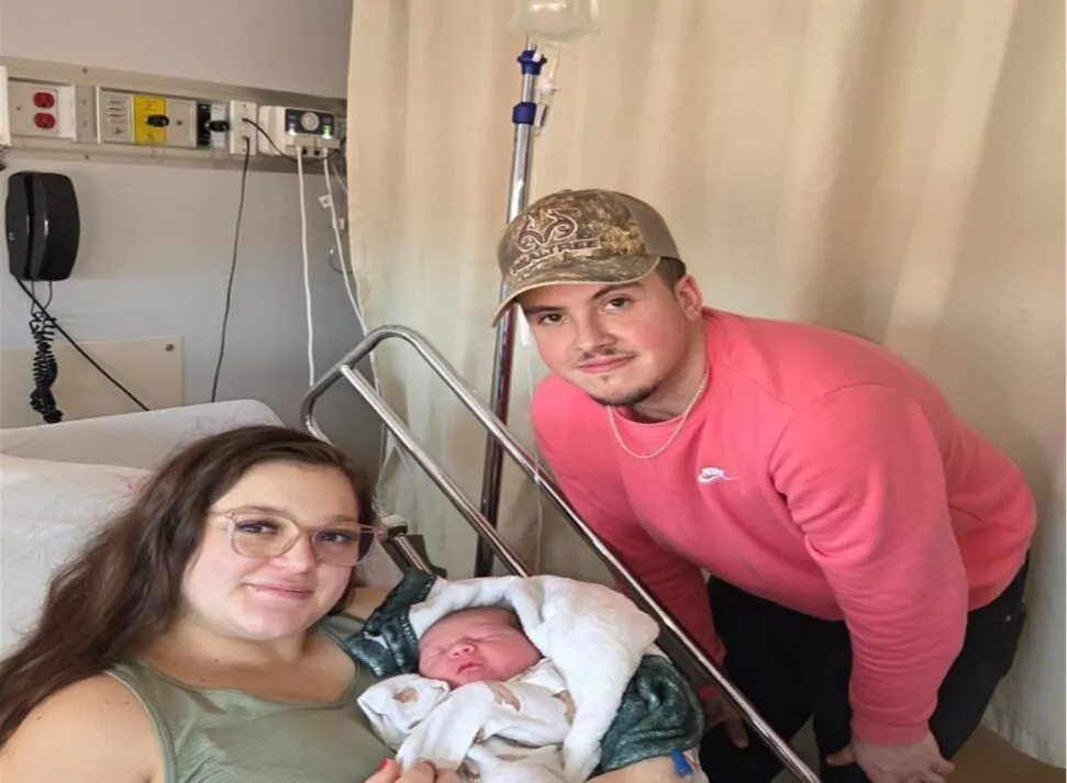 Une maman qui vient d'accoucher et qui est allongée dans un lit d'hôpital tient dans ses bras son nourrisson, alors que le papa se tient à côté.