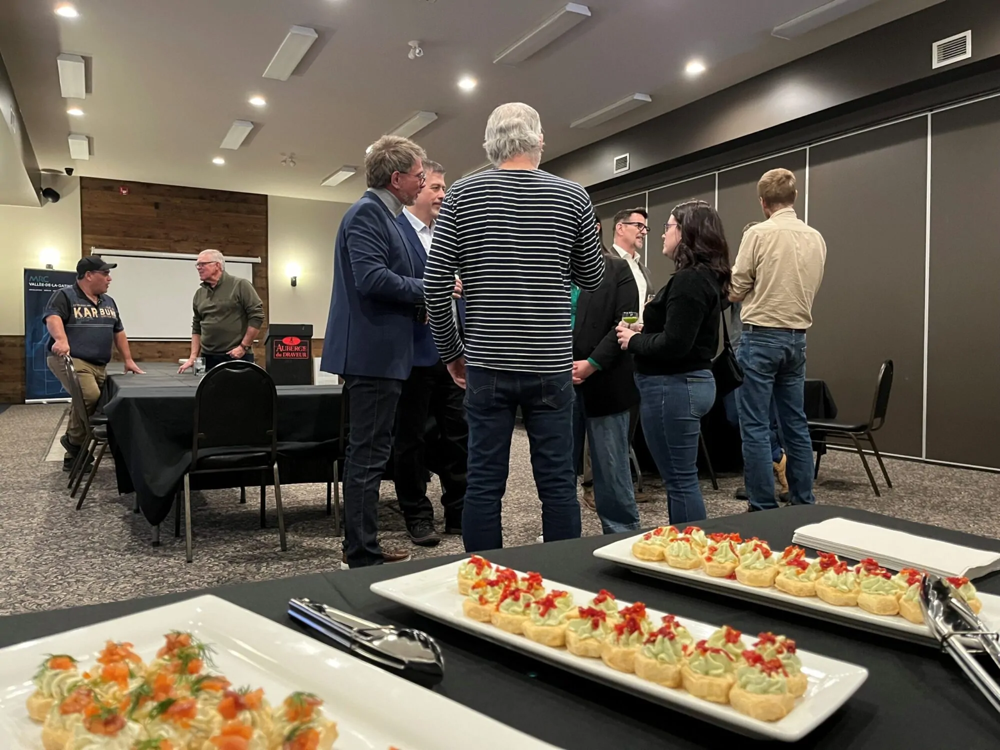 Dans une salle de réception aux murs griss, plusieurs adultes discutent en petits groupes autour de tables recouvertes de nappes noires. Au premier plan, sur une table, des plateaux de bouchées élégantes sont soigneusement disposés à côté de pinces de service.