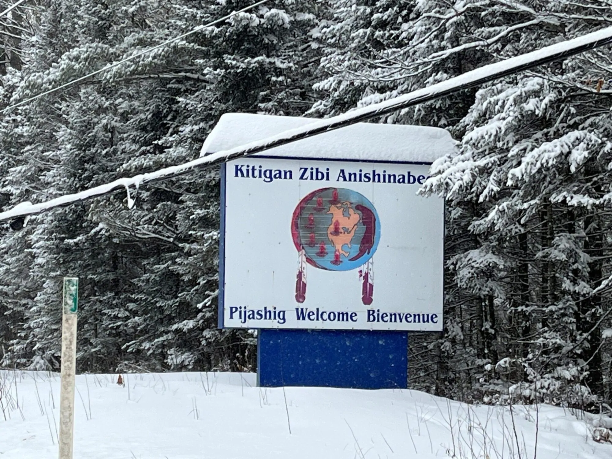 Un panneau bordé de bleu, couvert de neige, se dresse en bordure d’une route forestière enneigée. On y lit « Kitigan Zibi Anishinabeg » au-dessus d’un symbole circulaire représentant une carte et des plumes, ainsi que « Pijashig Welcome Bienvenue », entouré d’arbres chargés de neige et traversé par des fils électriques.