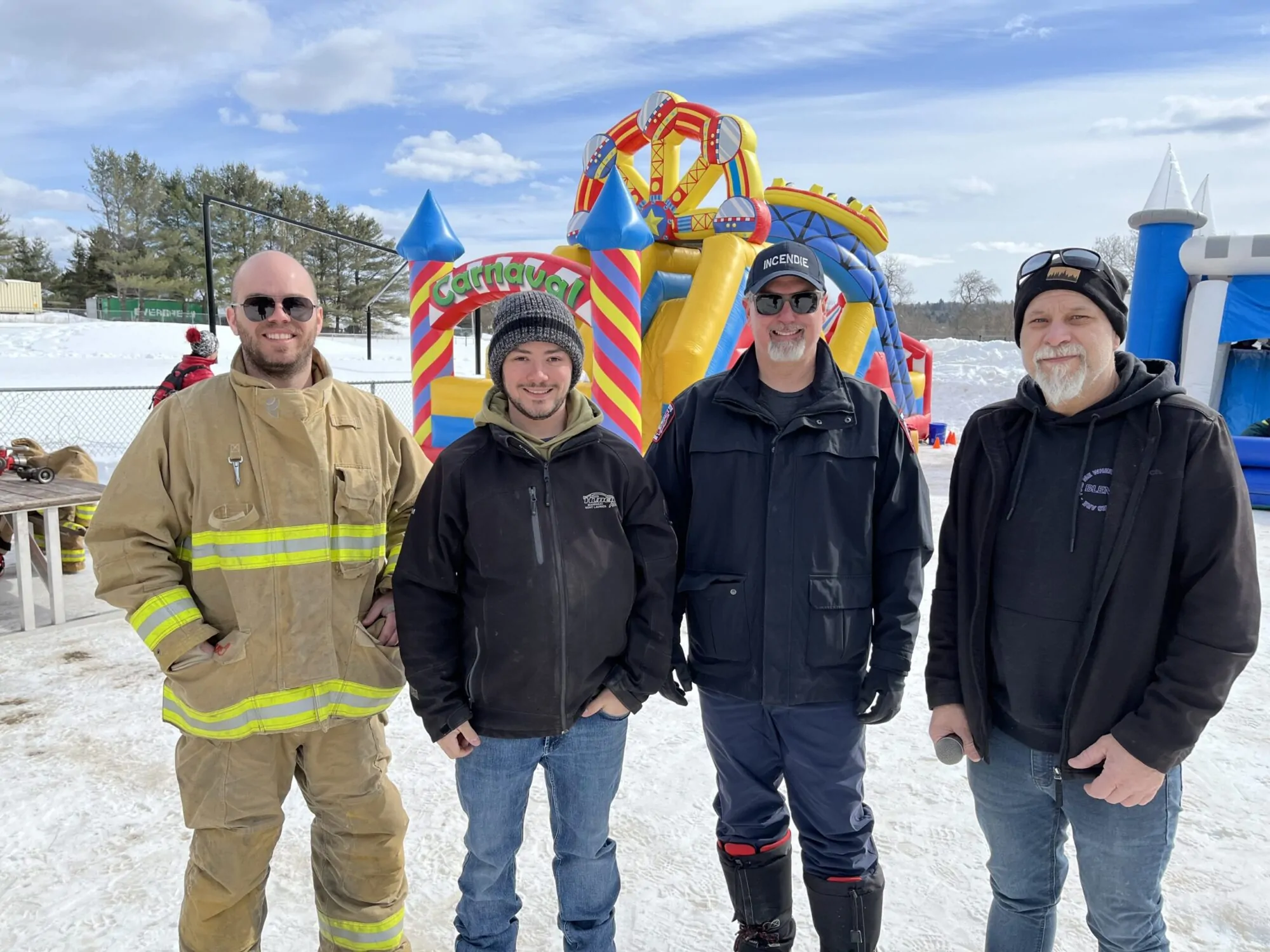 Quatre hommes, dont un avec son habit de pompier, sont rassemblés avec des structures de jeu gonflable derrière eux durant une belle journée d'hiver.