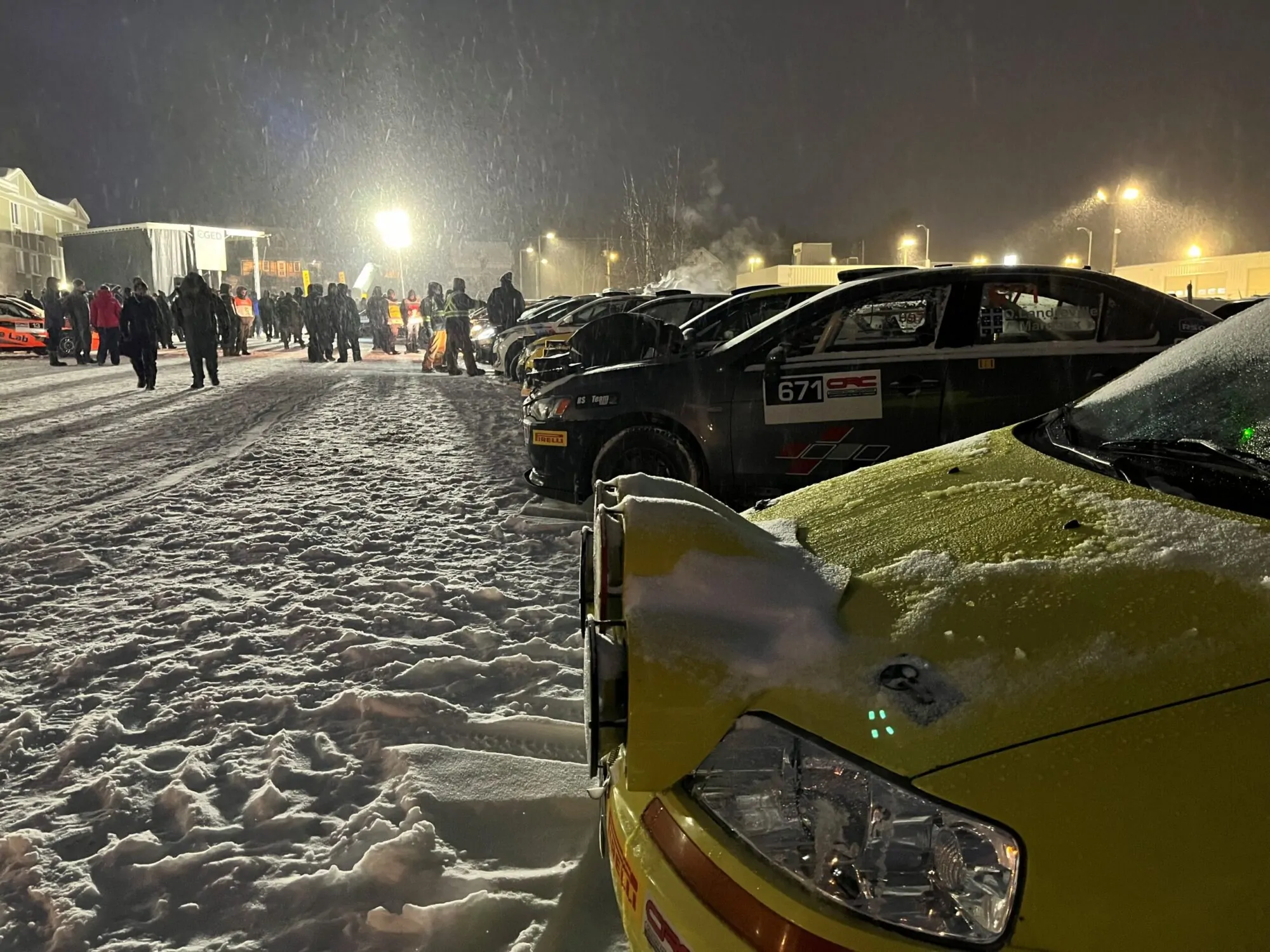 Une rangée de voitures de course est stationnée sur un site enneigé la nuit, éclairé par de puissants projecteurs. De nombreuses personnes en manteaux d’hiver circulent entre les véhicules malgré le froid.