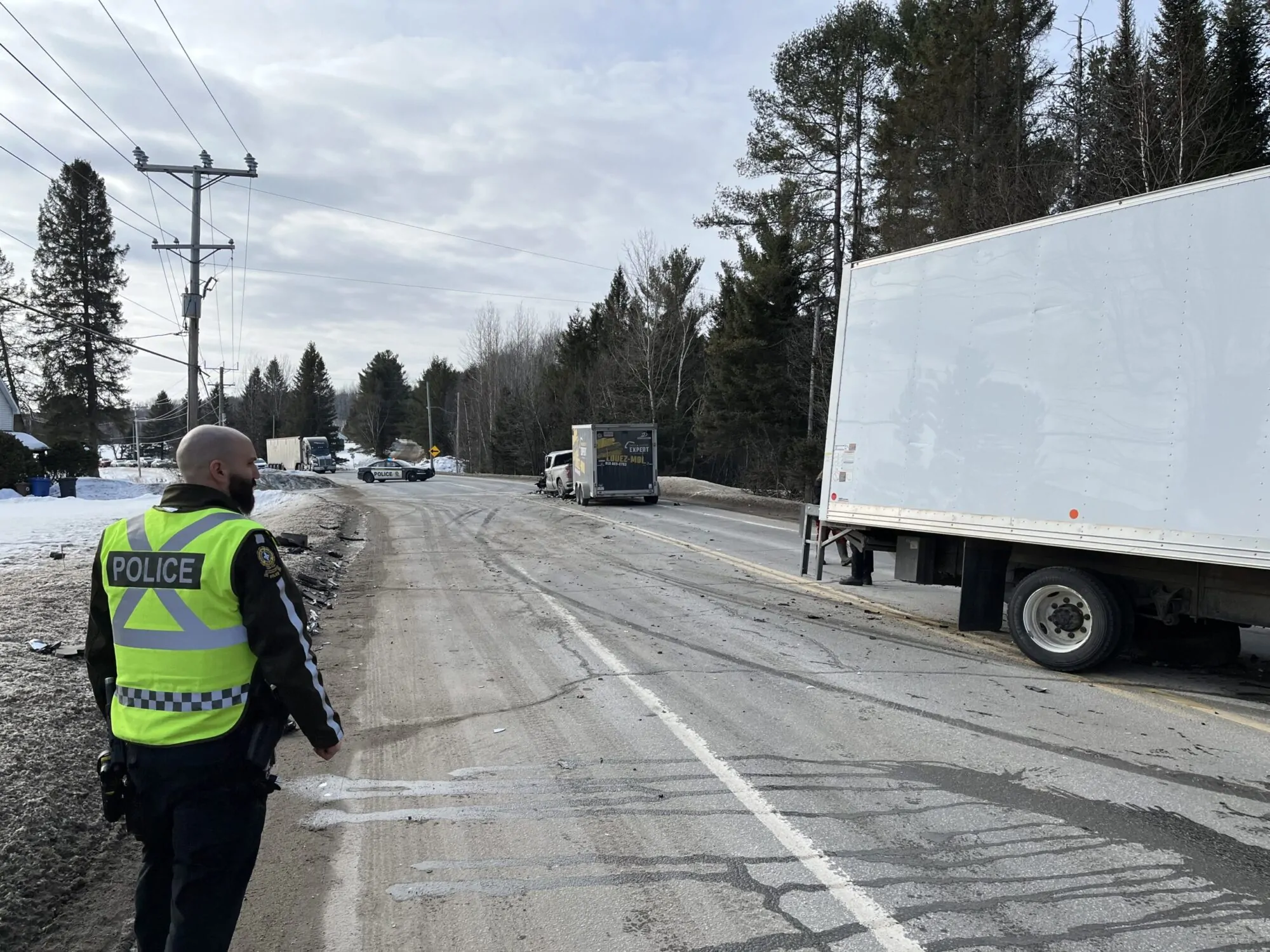 Un policier de la Sûreté du Québec portant un dossard jaune, vu de dos, regarde en direction d'un pick-up et d'une remorque immobilisés sur une route à la suite d'un accident en hiver. Des débris jonchent le sol près d'un camion. On voit plus loin une voiture de police qui bloque la circulation.